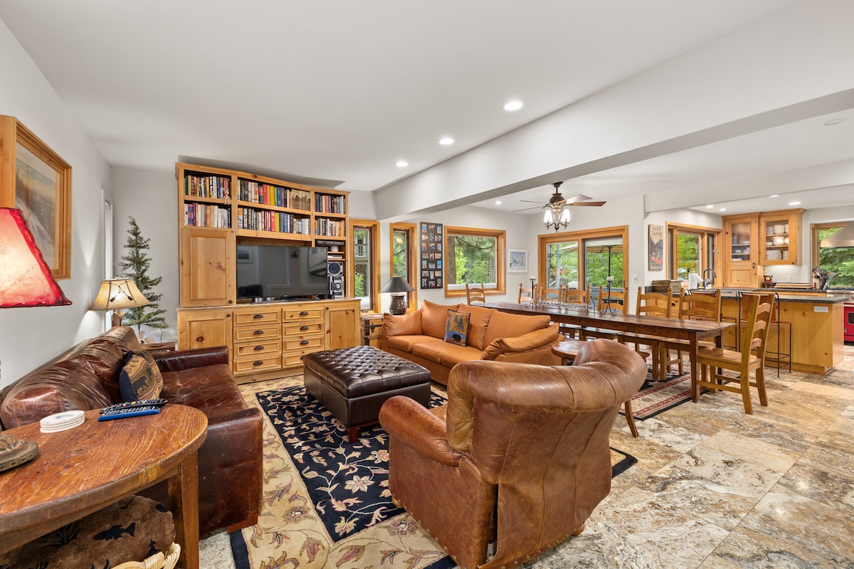 A spacious great room is furnished with comfortable seating arrangements, including a large leather sofa and armchairs. A dining table for 14 is positioned nearby, and natural light fills the area through multiple windows, providing views of the surrounding greenery.