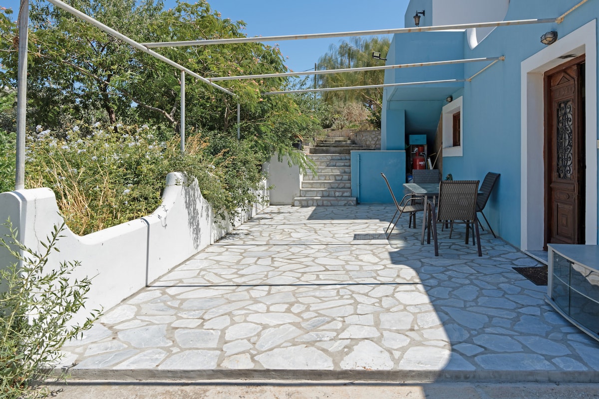 A spacious outdoor terrace is shown, featuring a stone-paved floor and a canopy for shade. A set of four dark wicker chairs surrounds a table against a backdrop of greenery. Steps lead up to a higher area, creating an inviting entryway to the property.