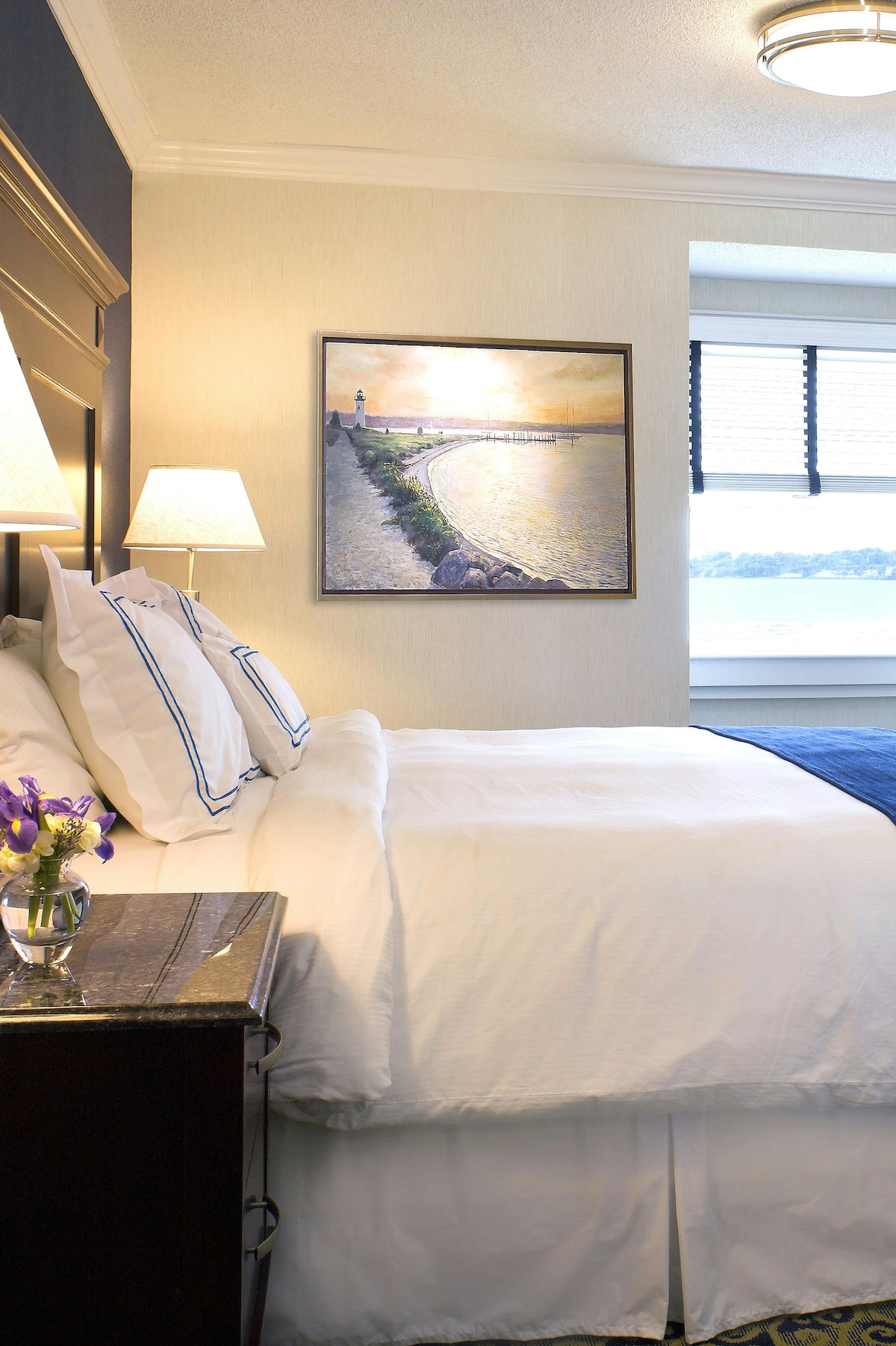 A well-appointed sleeping area features a spacious bed with crisp white linens and decorative blue accents. A framed artwork depicting a waterfront scene is displayed above a dark wooden nightstand, which holds a vase of fresh flowers. Natural light enters through a large window with black blinds.