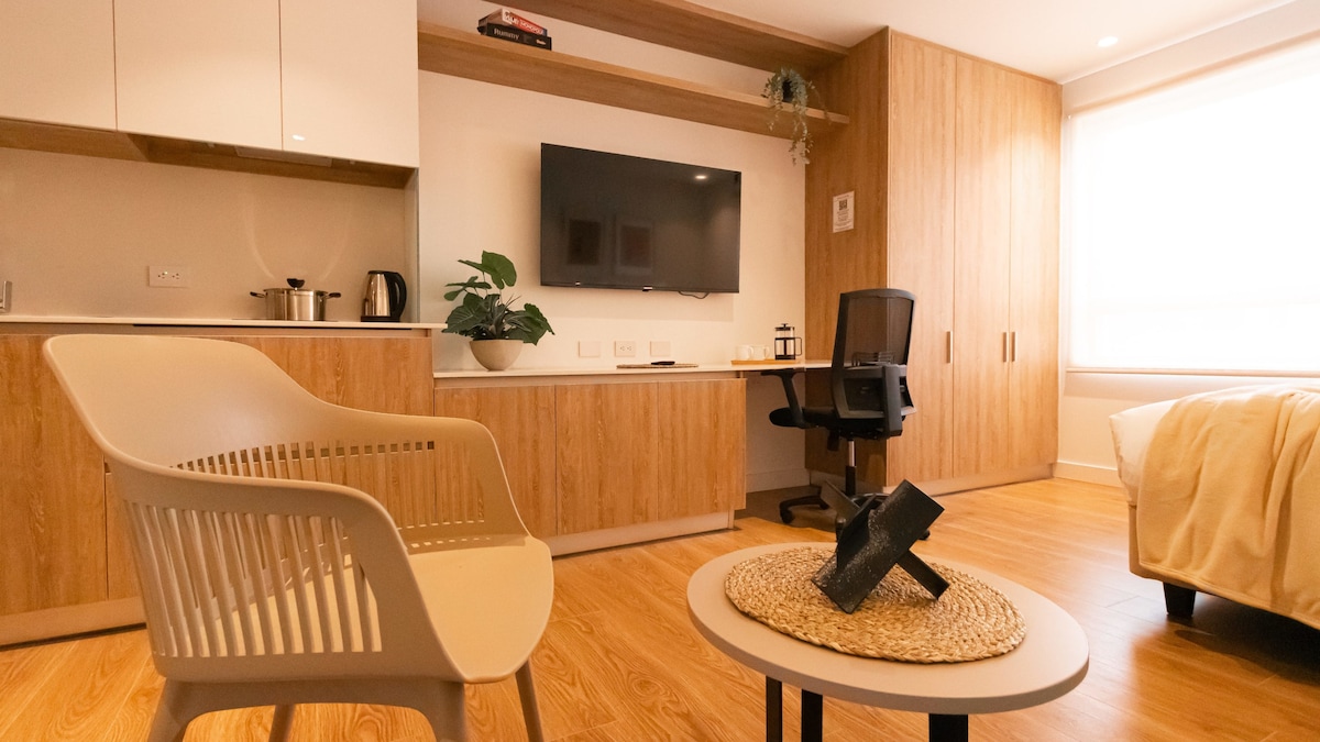 A modern living space features a comfortable chair with a minimalist design, situated beside a round table with a woven placemat. A sleek desk and an office chair are positioned opposite a wall-mounted Smart TV, with warm wood paneling adding a natural element to the decor.