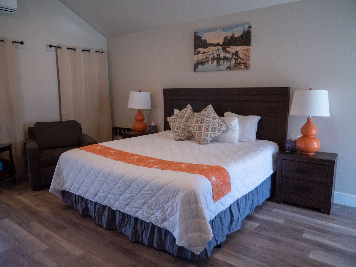 A spacious sleeping area features a king bed with a quilted white cover and decorative pillows. Two bedside lamps provide soft illumination. A cozy chair is positioned nearby, and natural light enters through a window adorned with simple curtains. Wall art adds a touch of character.