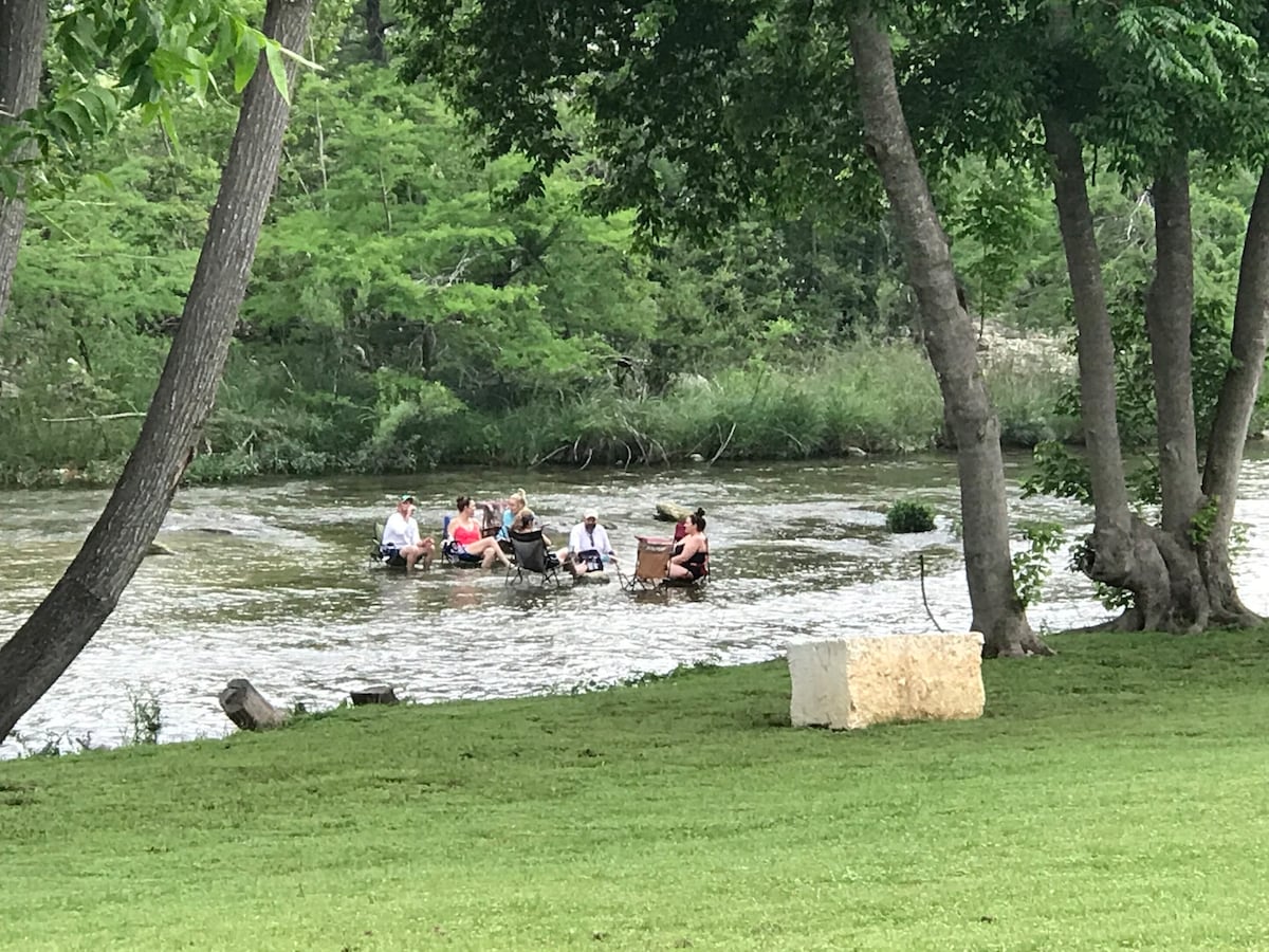 A natural setting features a grassy area near the Guadelupe River, where several individuals are seen enjoying a paddle activity. Surrounding trees provide shade, contributing to a serene outdoor environment.