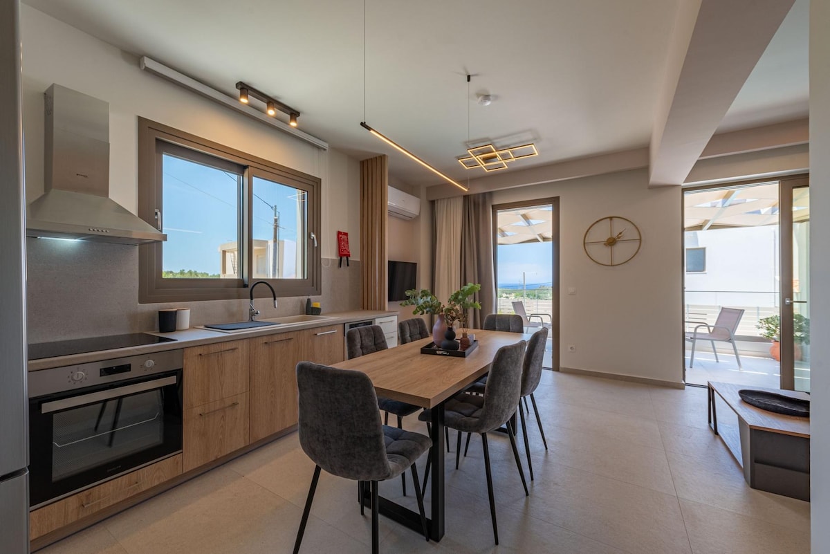 The open-plan kitchen and dining area features a modern design with wooden cabinetry and stainless steel appliances. A dining table with seating for six is placed centrally, while large windows allow light to fill the space, offering views of the outdoor terrace.
