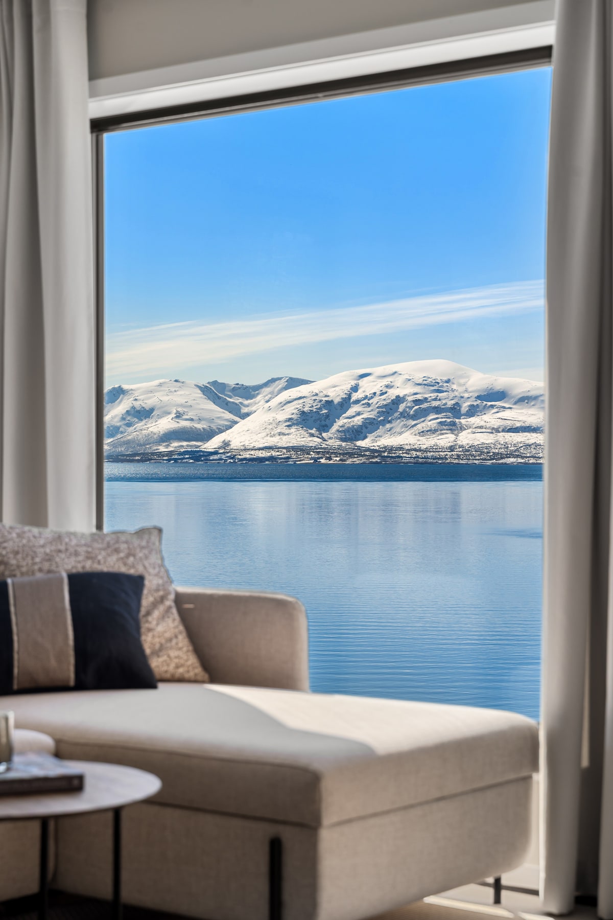 A large window offers a clear view of snow-capped mountains reflecting on calm water, with a cozy seating area in the foreground. Neutral-colored cushions adorn a modern sofa, enhancing the serene atmosphere.