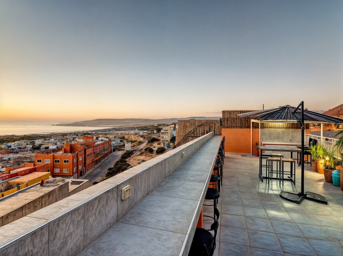A shared rooftop space features a long countertop with several stools, complemented by a dining table under a gazebo. The panoramic view of the coastline and village is showcased, as the sun sets, casting warm hues across the sky.