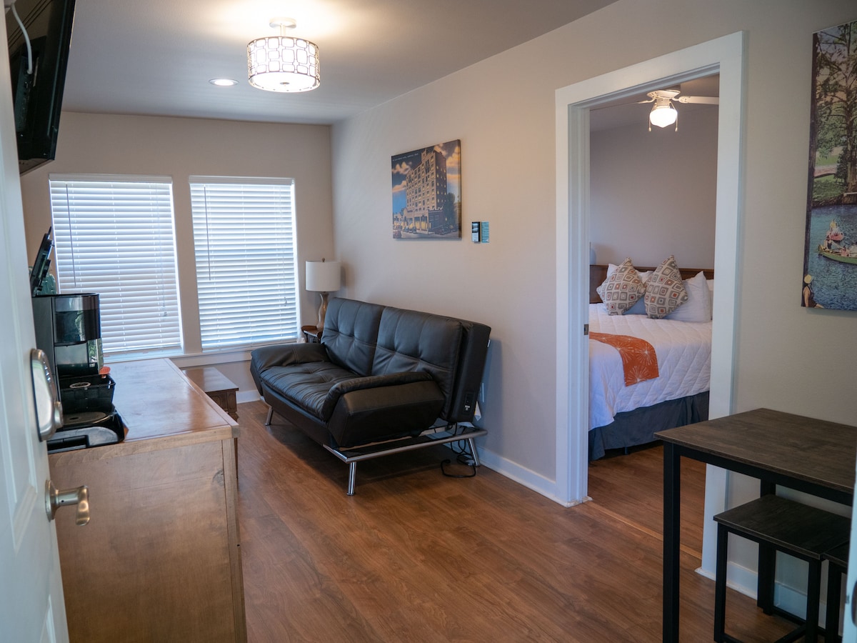 A spacious living area is featured with a fold-out couch facing a flat-screen TV. Natural light enters through two windows adorned with white blinds. A doorway leads to the bedroom, which showcases a king bed, complemented by patterned pillows and light-colored bedding.