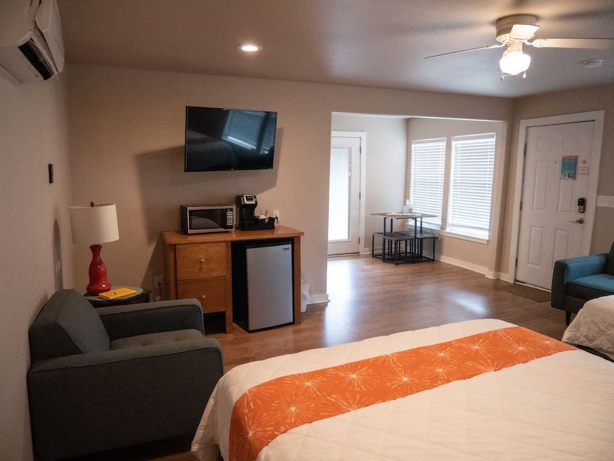 The spacious unit features two queen beds with a warm orange accent blanket. A kitchenette area with a microwave and mini fridge is present, alongside a flat-screen TV mounted on the wall. Natural light enters through the windows, highlighting a small dining table near the entrance.