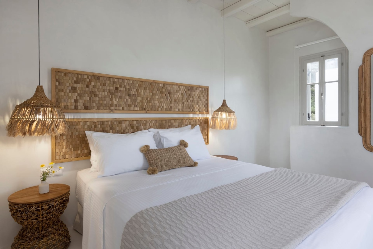 A serene bedroom features a spacious bed adorned with crisp white linens and a textured throw. Two woven pendant lights provide soft illumination beside the bed, which is flanked by round side tables. A small window offers natural light, enhancing the minimalist decor.