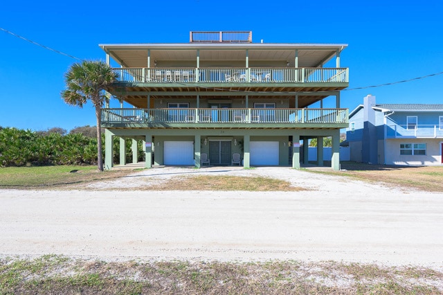The multi-level oceanfront estate is showcased against a clear blue sky. The property features expansive wrap-around balconies on each level, providing ample outdoor space. Below, the ground floor includes garages framed by a sandy path leading to lush greenery on either side.