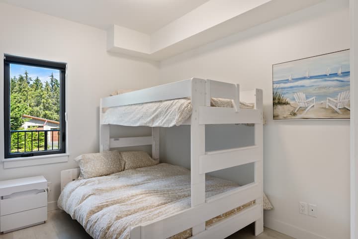 Útulná spálňa s bielou poschodovou posteľou a vzorovanou posteľnou bielizňou. Nočný stolík stojí vedľa okna s výhľadom na bujnú zeleň. Steny sú zdobené pokojnou maľbou plážových stoličiek. Ideálne pre rodiny alebo skupiny, ktoré navštevujú Bacoor.