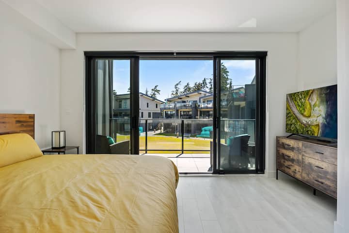 Svetlá spálňa s pohodlnou posteľou. Vychutnajte si prístup na vlastný balkón cez posuvné sklenené dvere, ktoré ponúkajú výhľad na okolitý pozemok. Vo vnútri je drevená komoda s televízorom. Vedľa postele je nočný stolík s lampičkou.