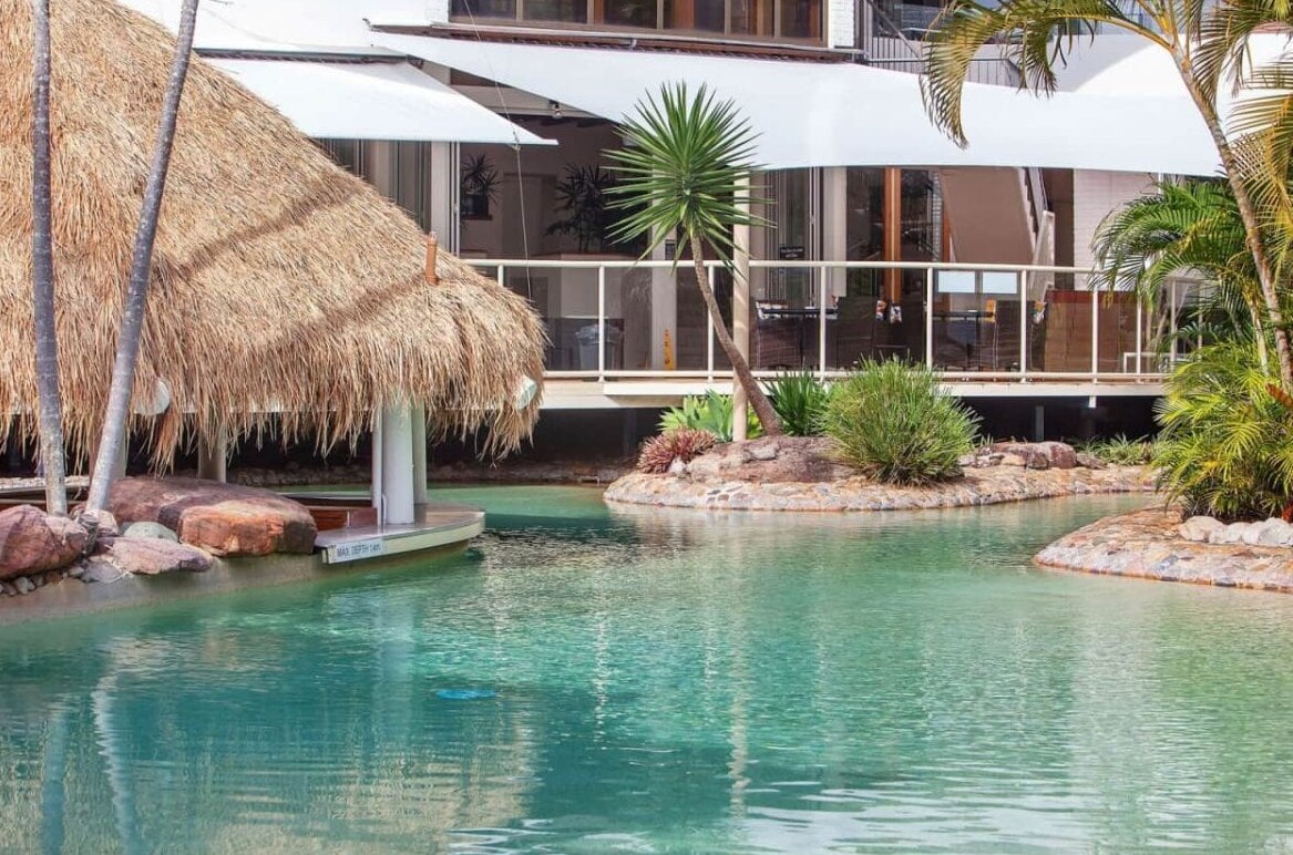 A tranquil view of a resort pool surrounded by lush greenery and natural rock formations. Thatched-roof cabanas provide shade near the water's edge, reflecting a serene atmosphere. The clear water showcases shades of turquoise, inviting relaxation and leisure.