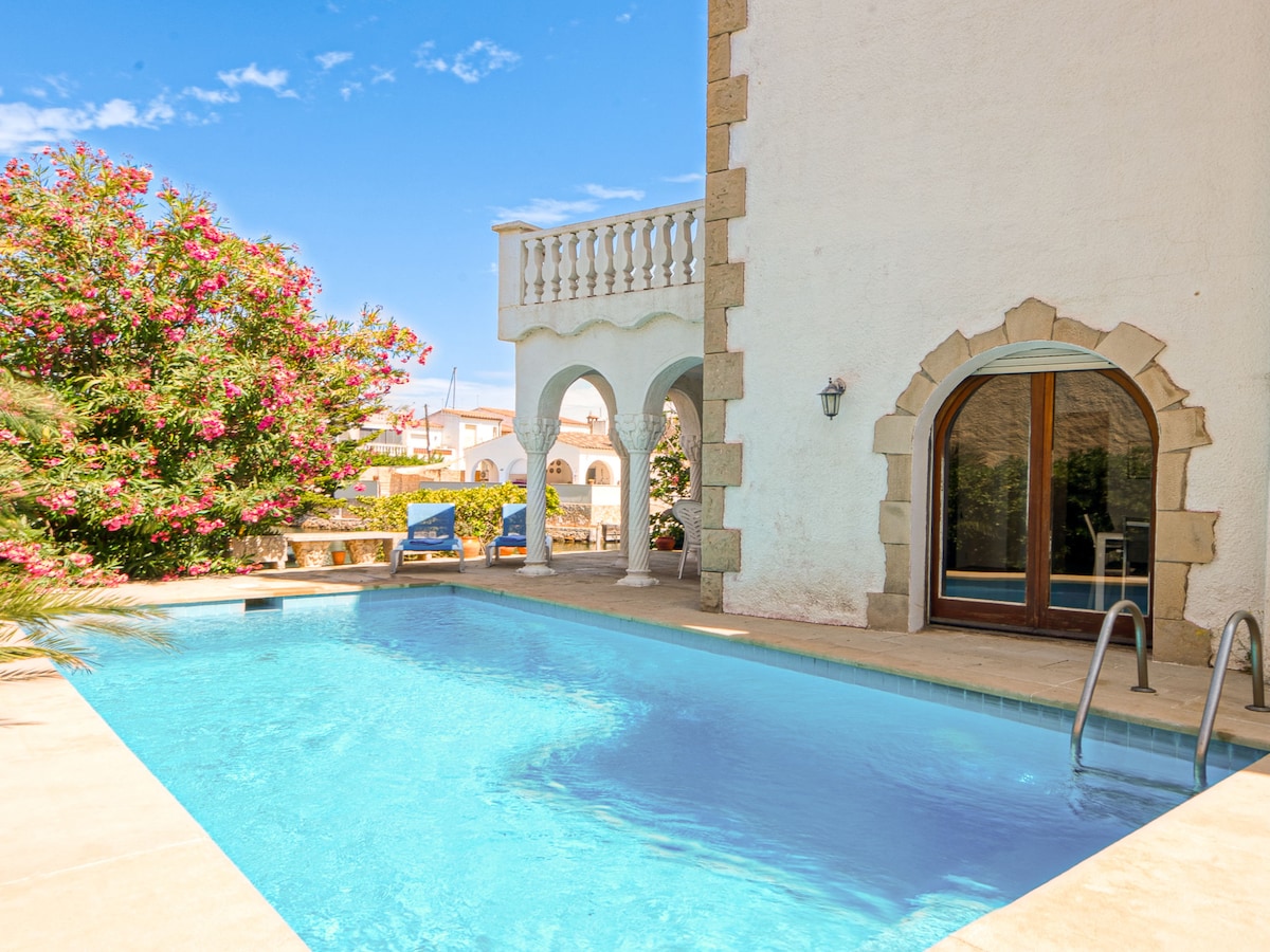 The image captures an inviting swimming pool bordered by a stone terrace. Lush greenery and flowering plants frame the area, while the house's distinct architectural features, including an arched doorway and stone detailing, provide a charming backdrop under a clear blue sky.