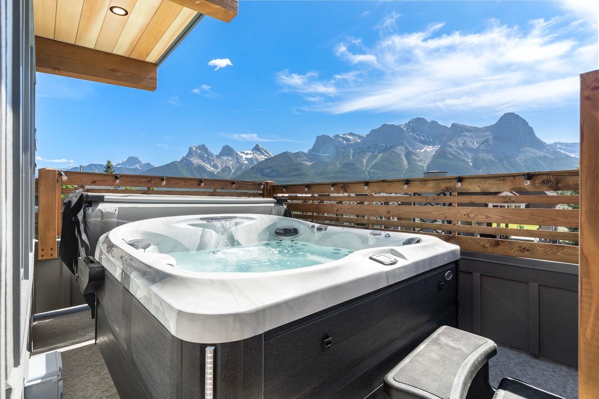 A private hot tub is positioned on a spacious deck, offering sweeping views of the surrounding mountains. Sunlight glistens on the water's surface, while wooden railings provide a sense of privacy and warmth to the outdoor space.
