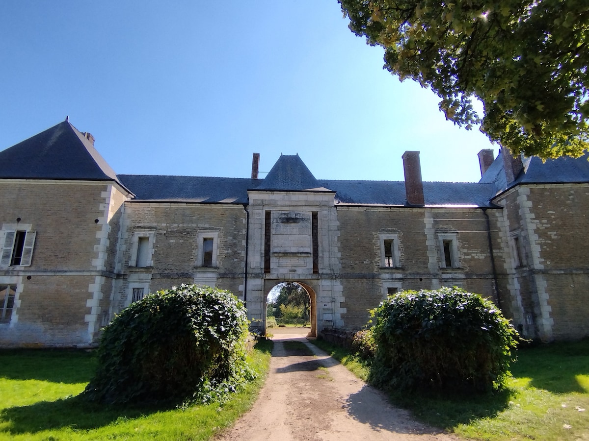 The image showcases the exterior of a historic château, featuring a central archway connecting two sections of the building. Lush greenery frames the entrance, with a dirt path leading into the estate. A clear blue sky complements the stone facade and slate roofs.