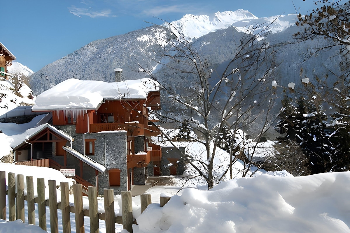 A charming chalet is nestled among snow-covered landscapes, with a wooden fence framing the view. The mountains rise majestically in the background, while the clear blue sky enhances the serene winter atmosphere surrounding the warm-hued buildings.