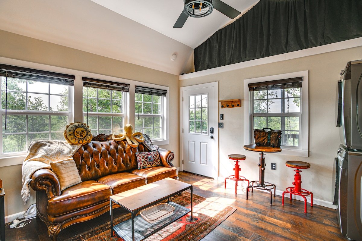 A cozy living area features a leather sofa with decorative pillows, positioned near large windows that allow natural light to fill the space. A wooden coffee table rests on a textured rug, and two tall stools provide seating at a small side table. A ceiling fan is installed above.