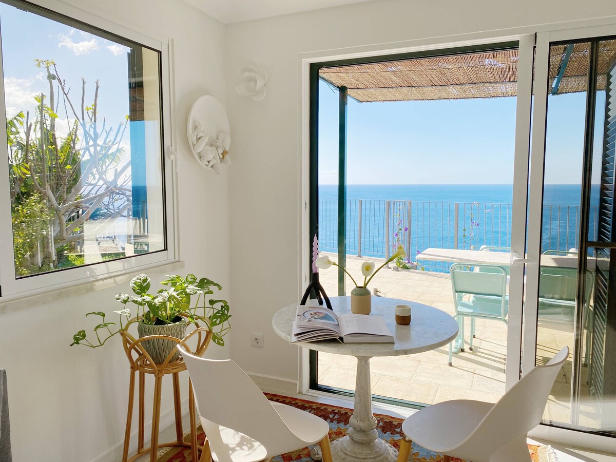 A bright and airy dining area is highlighted by a round table with a light-colored surface. Two modern chairs frame the table, while a plant adds a touch of greenery. Large glass doors provide a panoramic view of the ocean beyond the terrace.