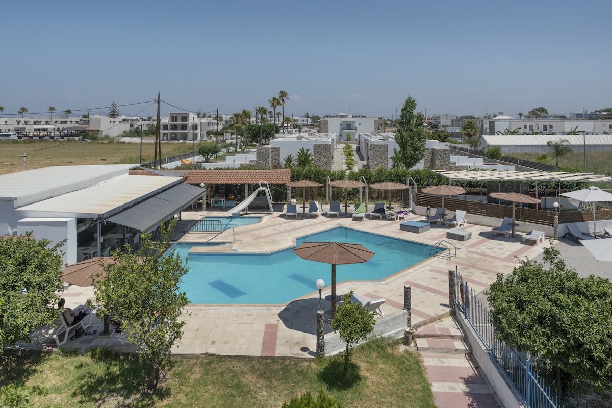 An expansive outdoor pool area features a large swimming pool surrounded by sun loungers and shaded seating under umbrellas. Lush greenery is visible along the perimeter, with well-maintained pathways leading to various areas, creating a spacious atmosphere for relaxation.