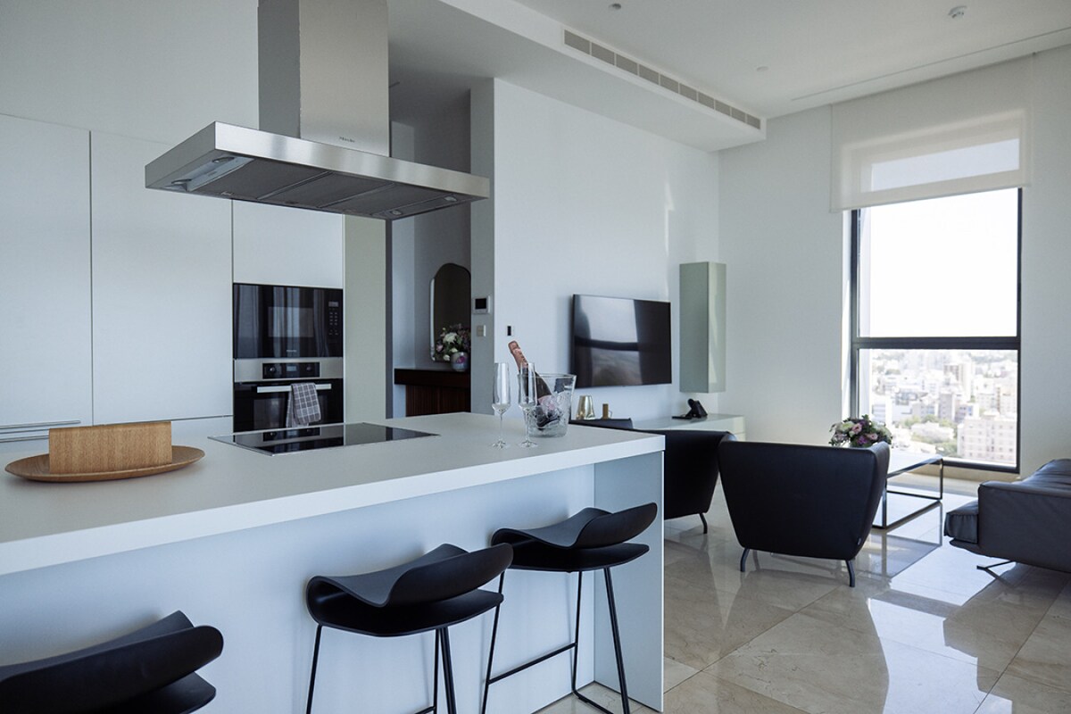 A modern kitchen and living area are featured, with sleek cabinetry and a central island. Contemporary bar stools are arranged around the island, while a flat-screen television is mounted on the wall. Large windows provide natural light and a view of the city.