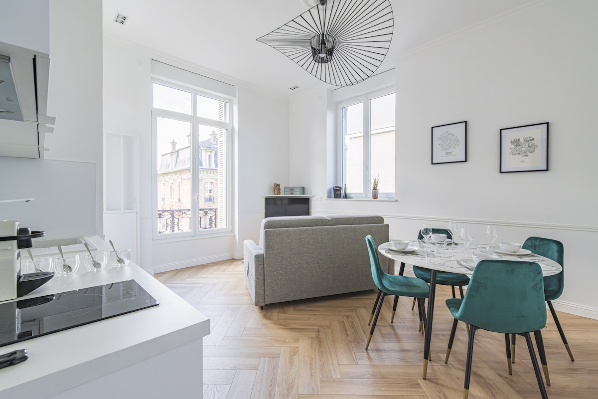 An open living and dining area features large windows allowing natural light to fill the space. A comfortable sofa is positioned near a modern dining table set with elegant dishware. The light hardwood floor complements the minimalist décor, creating a welcoming and airy atmosphere.