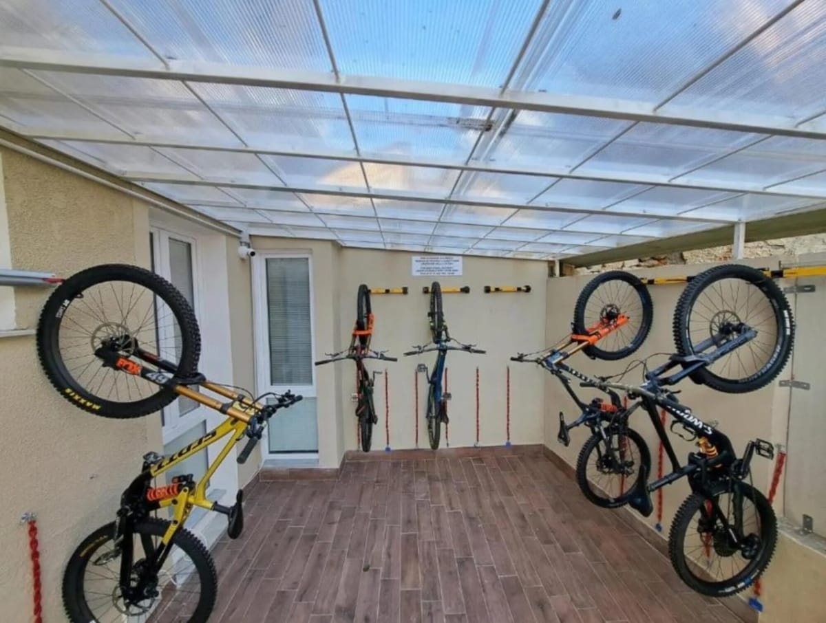 A covered storage area features several bicycles securely mounted on wall racks. A tiled floor enhances functionality, while natural light filters through the transparent roofing, illuminating the space. The entrance door is visible, providing easy access.