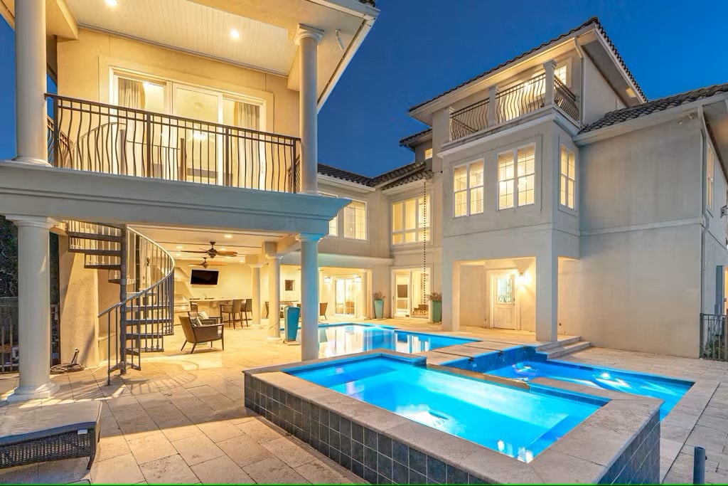 The outdoor area of the villa showcases a private pool and spa, surrounded by a spacious patio. A spiral staircase leads to a second-floor balcony, with multiple seating areas visible through expansive glass doors, allowing for a seamless connection to the indoor living spaces.