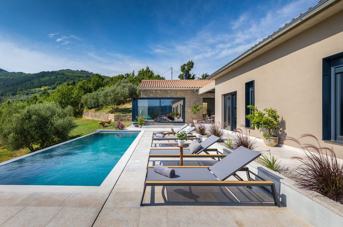 An outdoor area features a private pool with a spacious deck lined by sun loungers. Surrounding greenery enhances the tranquil setting, while large glass doors invite natural light into the villa. The sleek design and neutral colors contribute to an elegant atmosphere.