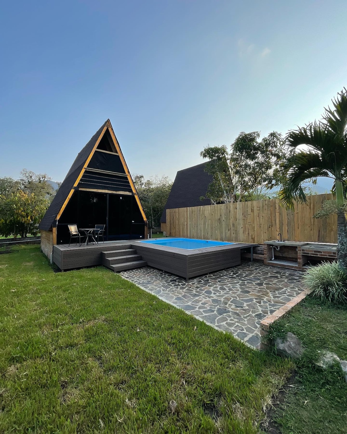 A charming cabin with a distinctive triangular roof is complemented by a private pool area. Sun loungers are positioned beside the pool on a wood deck, surrounded by a stone path. Lush greenery and wooden fencing enhance the outdoor ambiance, providing a sense of tranquility.