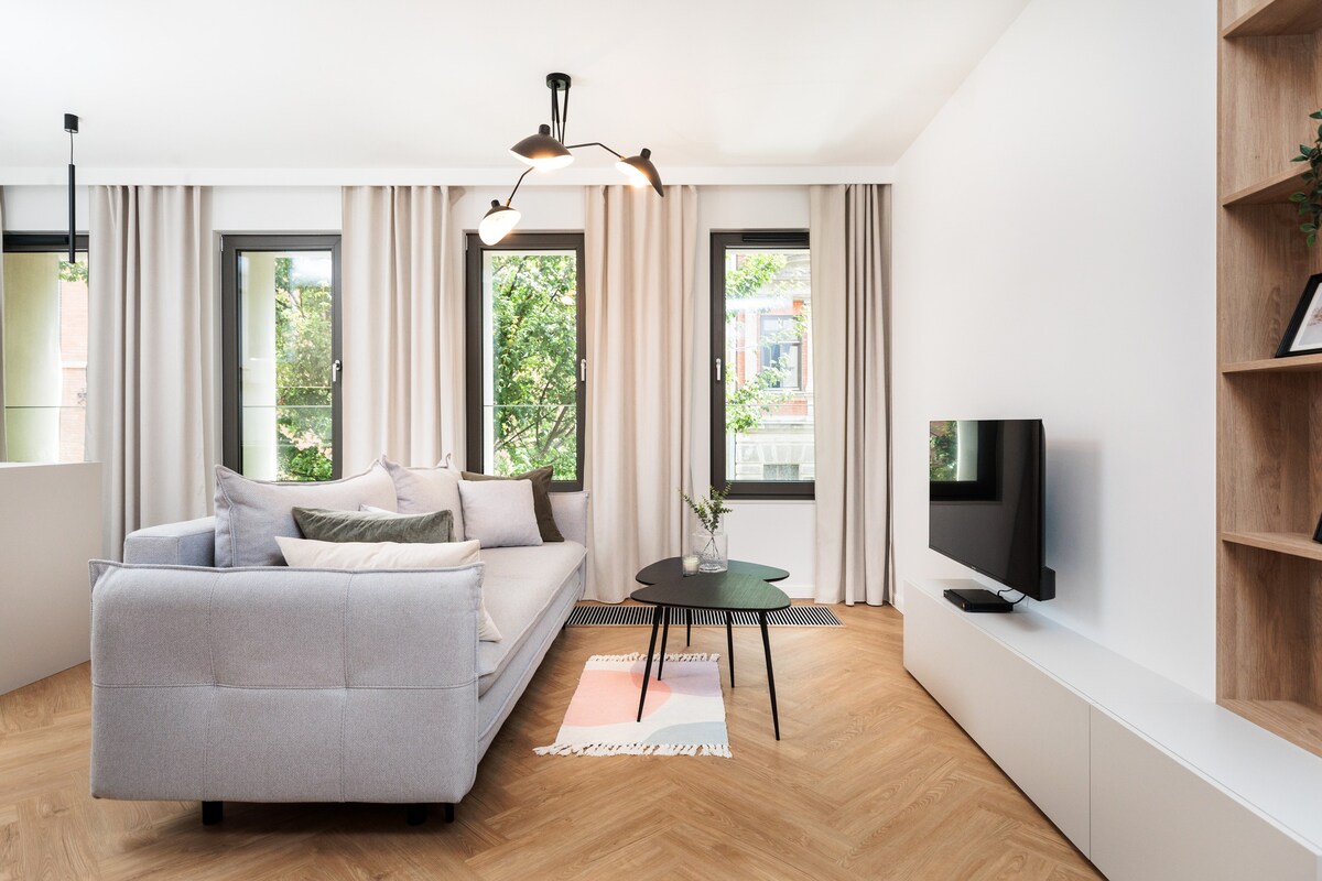A bright living room is featured, with a light gray sofa adorned with soft cushions. A round black coffee table sits centrally, while a television is mounted on the wall. Large windows with sheer curtains allow natural light to illuminate the space, surrounded by greenery.