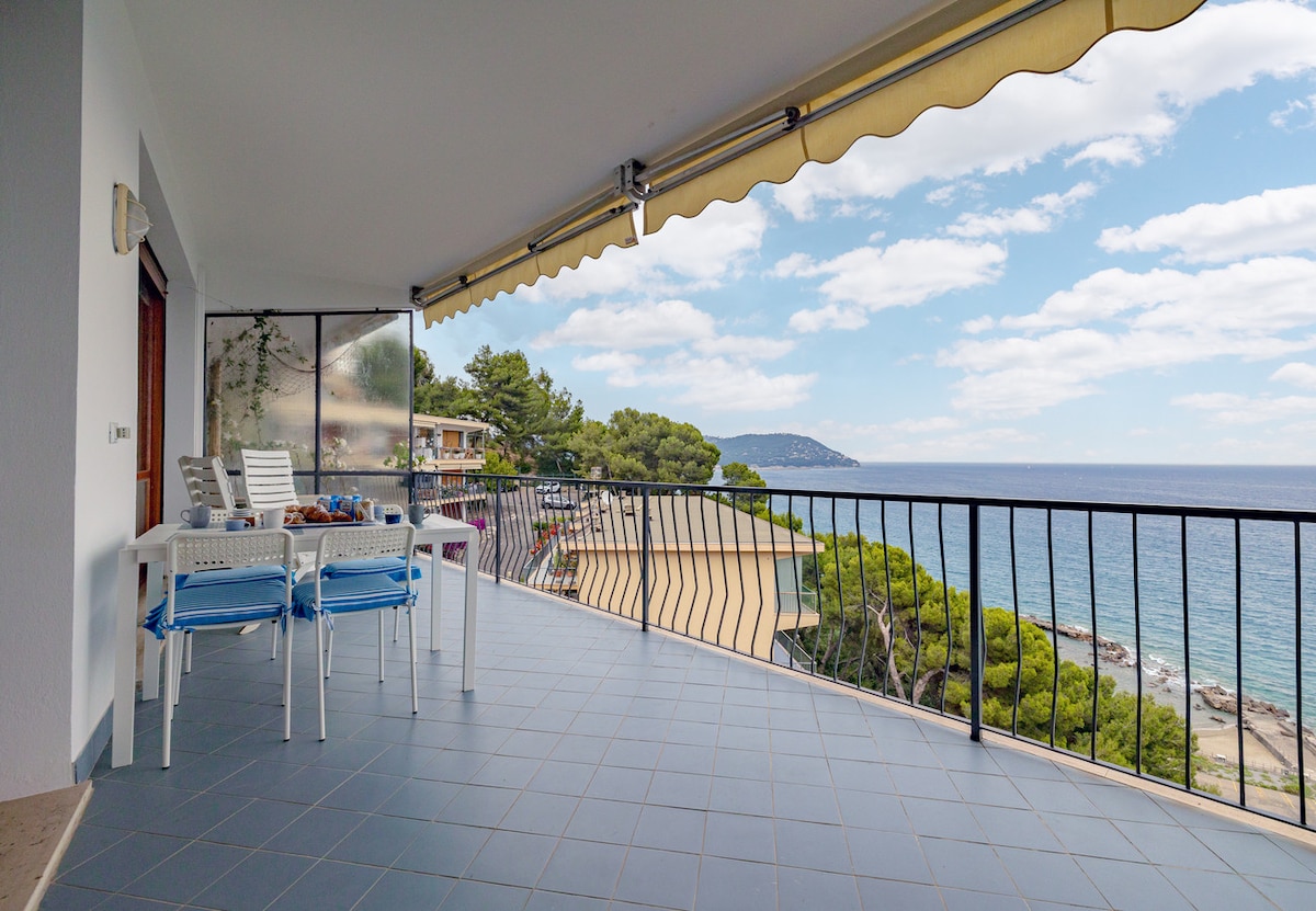 A spacious terrace is showcased, featuring a dining table set for meals. The expansive sea view stretches toward the horizon, framed by lush greenery. The soft curves of the coastline are visible, while a gentle cloud cover adds depth to the blue sky.