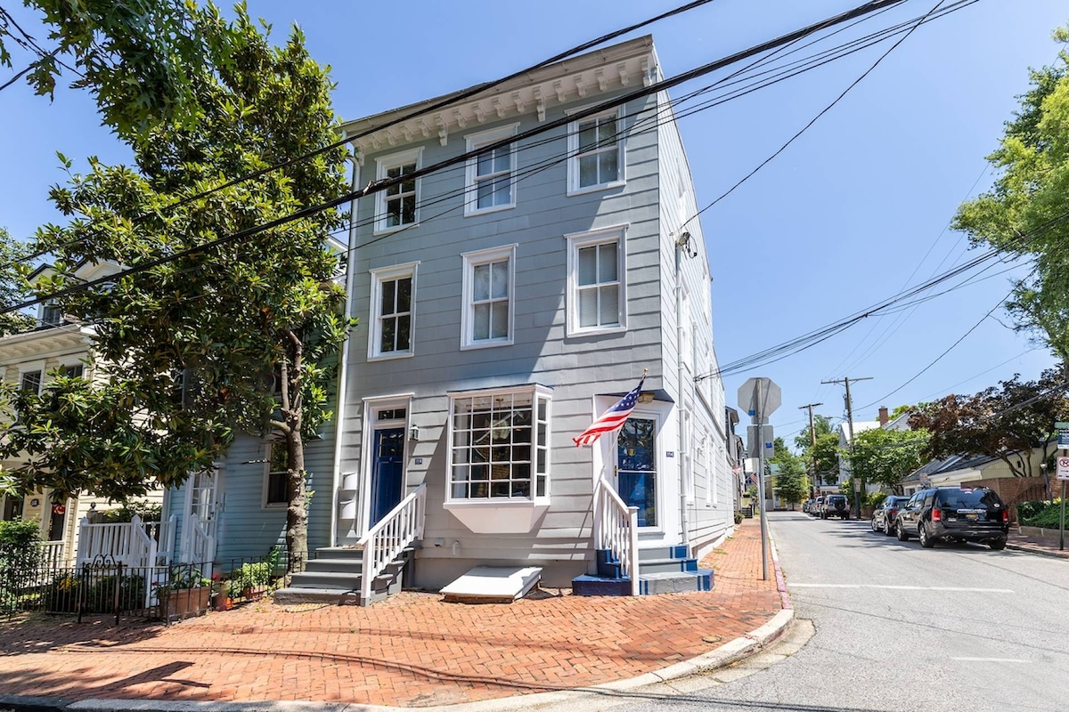 An exterior view of a three-story historic building is displayed, featuring a light blue facade and white trim. Steps lead to the front entrance, which is framed by large windows. A flag is proudly displayed on the porch, and the surrounding area includes brick walkways and greenery.