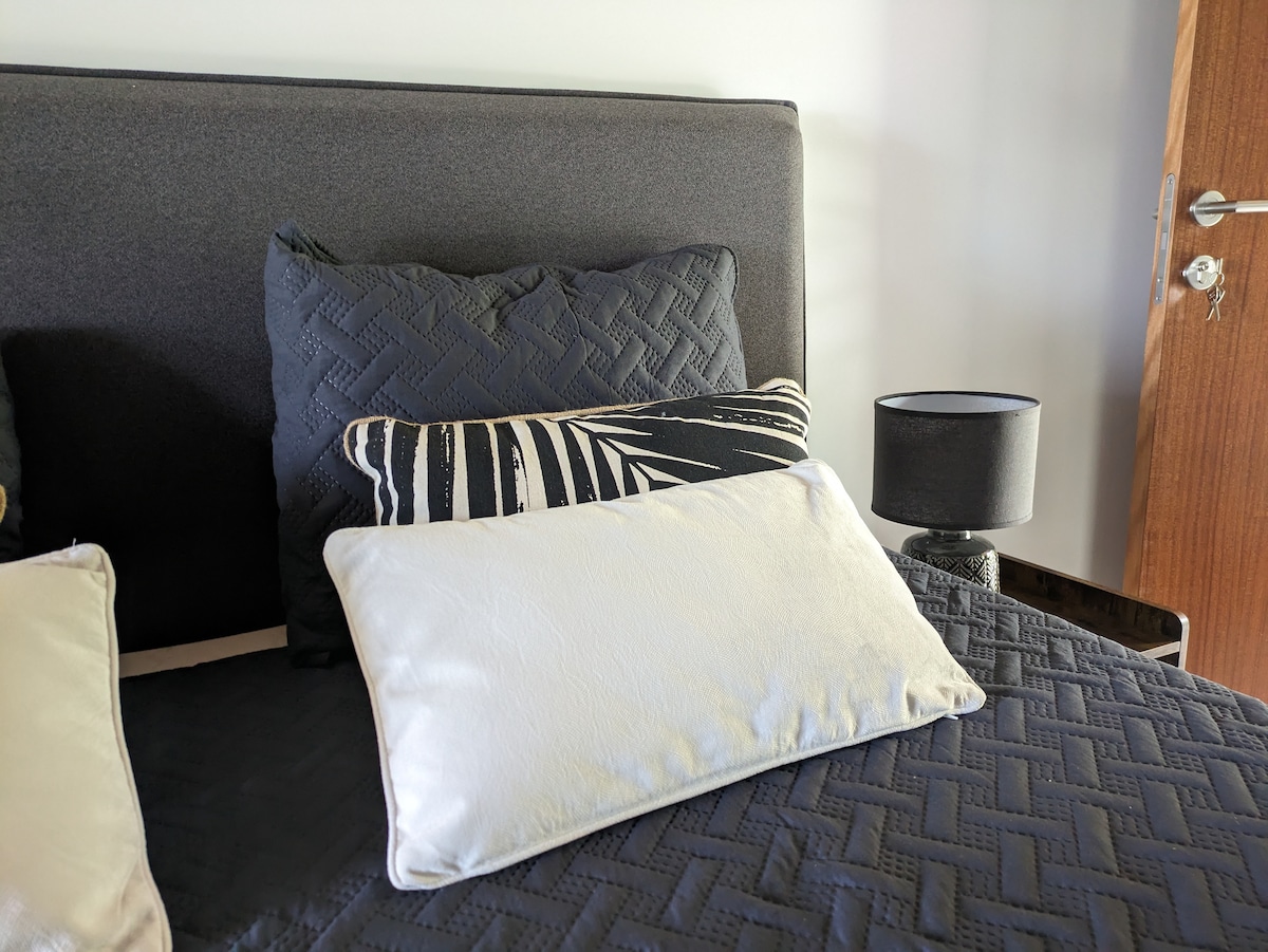 The queen size bed features a dark upholstered headboard adorned with a variety of decorative pillows, including a textured black option and a patterned one. A bedside lamp is situated next to the bed, providing a soft illumination against a neutral wall background.