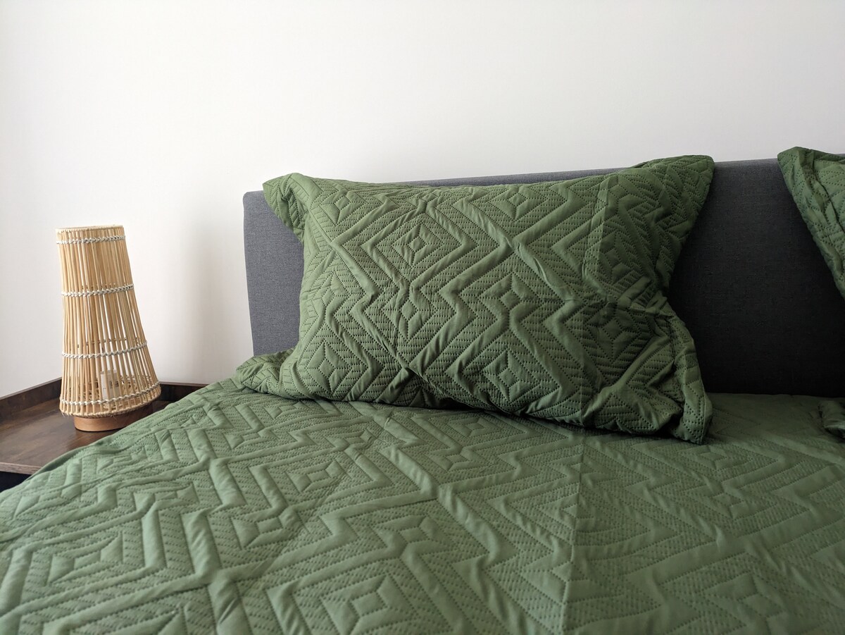 A queen size bed is dressed in a green quilt with a geometric pattern, complemented by coordinating green pillows. A woven lamp stands on the bedside table, accentuating the room's calming design and inviting ambiance.