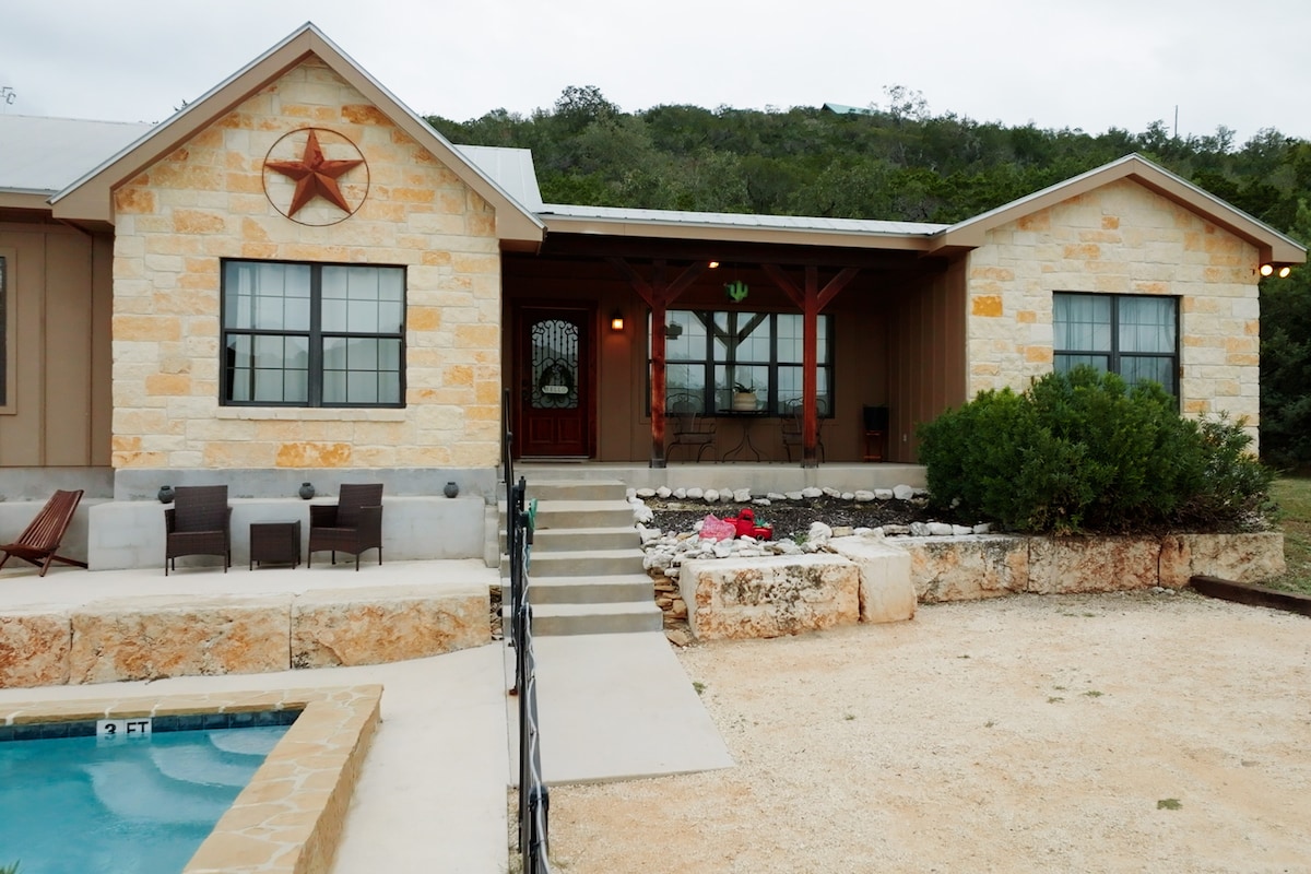 A charming exterior of the home features light-colored stone and a prominent star emblem above the entrance. A small staircase leads to a covered porch with seating, while landscaped stones and flowers adorn the area. A private pool edge can be seen to the left.