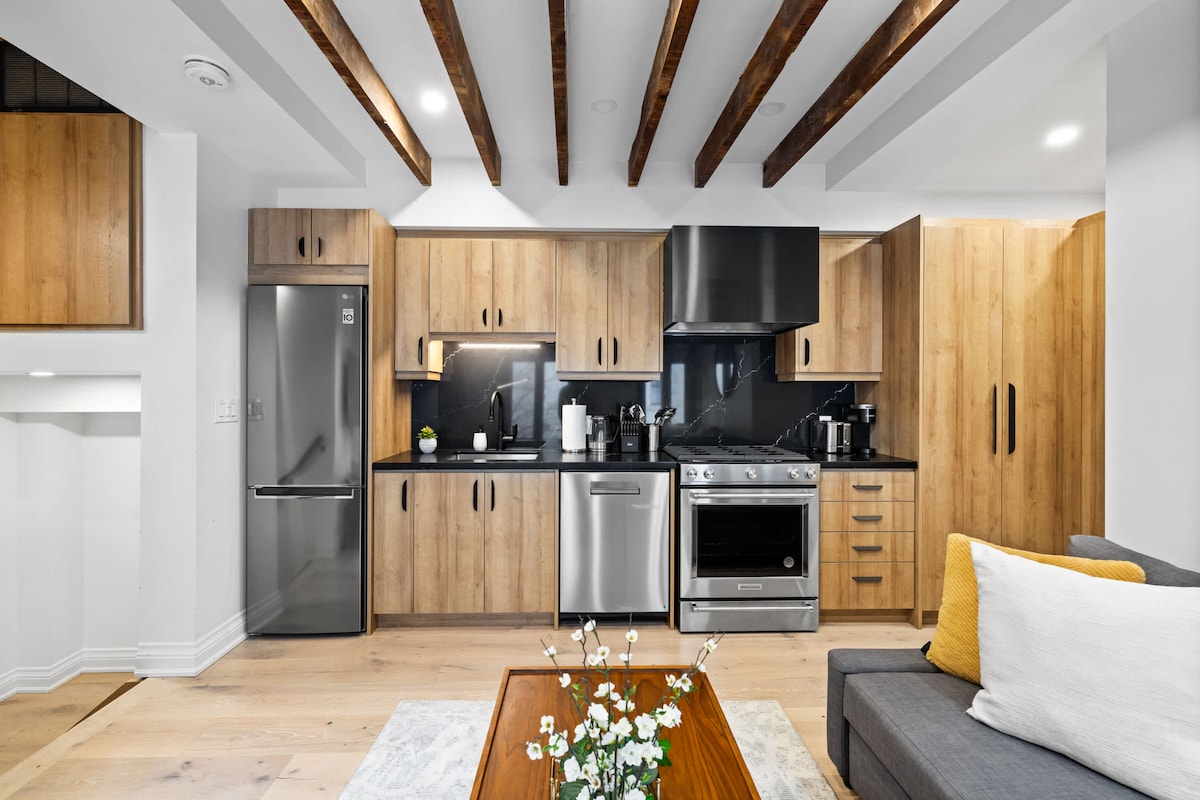 The modern kitchen features wood cabinetry paired with sleek black countertops. Stainless steel appliances, including a refrigerator, oven, and dishwasher, are highlighted. A cozy living area is visible in the foreground, furnished with a gray sofa and a wooden coffee table adorned with fresh flowers.