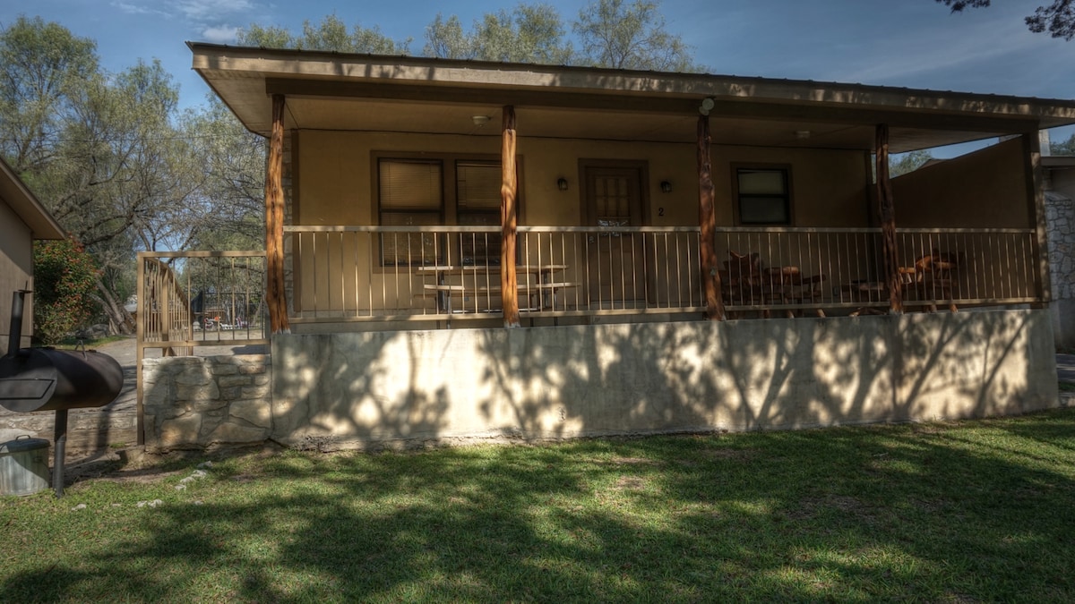 A spacious cabin is featured, showcasing a covered porch with wooden railings and several outdoor seating options. The structure is set against a backdrop of lush green grass and scattered trees, providing a serene environment.