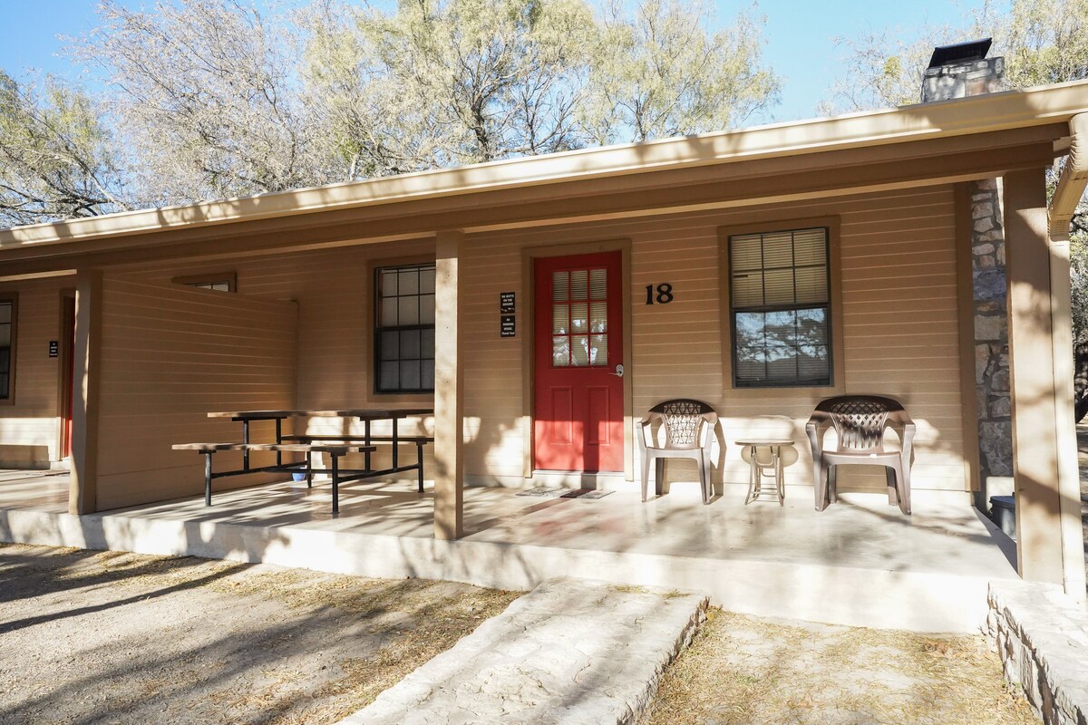 The cabin exterior features a covered porch with a picnic table and two chairs. A bright red door stands out against the light brown siding. Windows are framed in dark, complemented by a natural stone chimney next to the entrance.