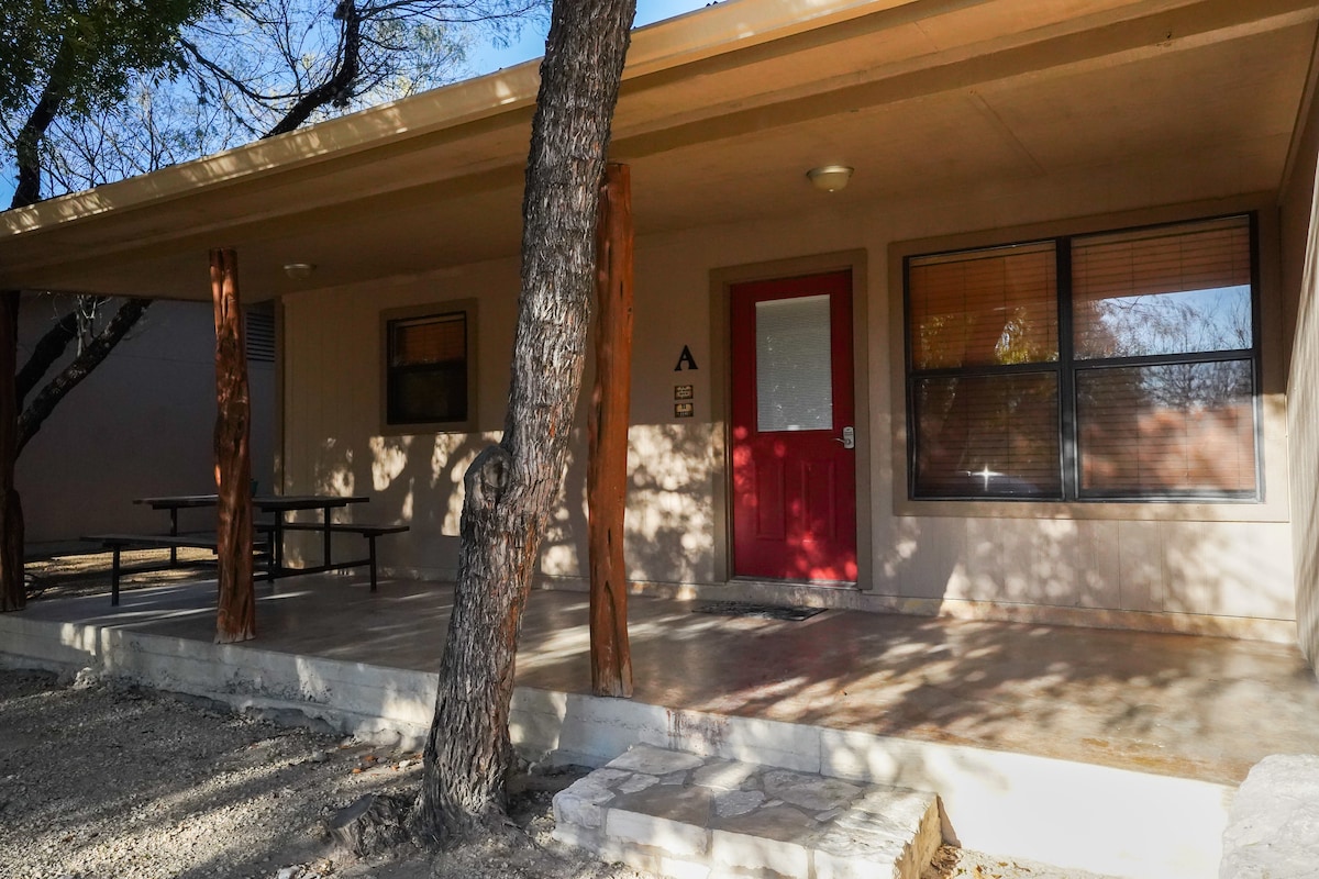 The entrance features a covered porch with natural wood pillars and a concrete floor. A bright red door is prominent, accompanied by multiple windows that allow natural light to fill the space. A picnic table is situated nearby for outdoor dining.