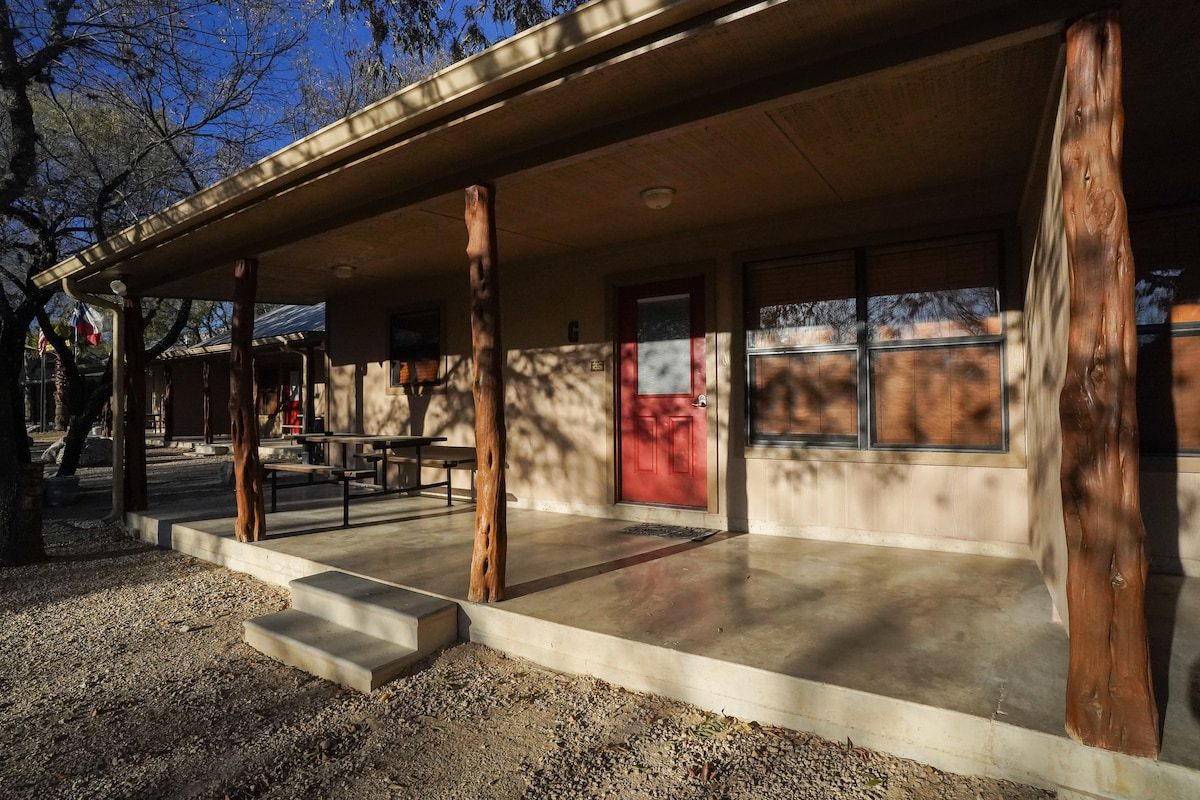 The cabin's exterior is presented with a covered porch supported by wooden pillars. A red door is highlighted at the entrance, with a concrete patio visible for outdoor gatherings. Gravel landscaping surrounds the cabin, enhancing the natural setting.