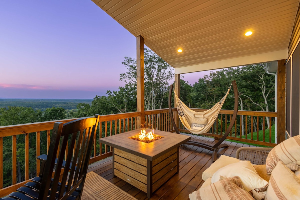 A spacious deck is featured, equipped with a fire pit surrounded by a wooden table. Comfortable seating includes a swinging hammock and rocking chairs, inviting relaxation while taking in the expansive view of the landscape and horizon at dusk.