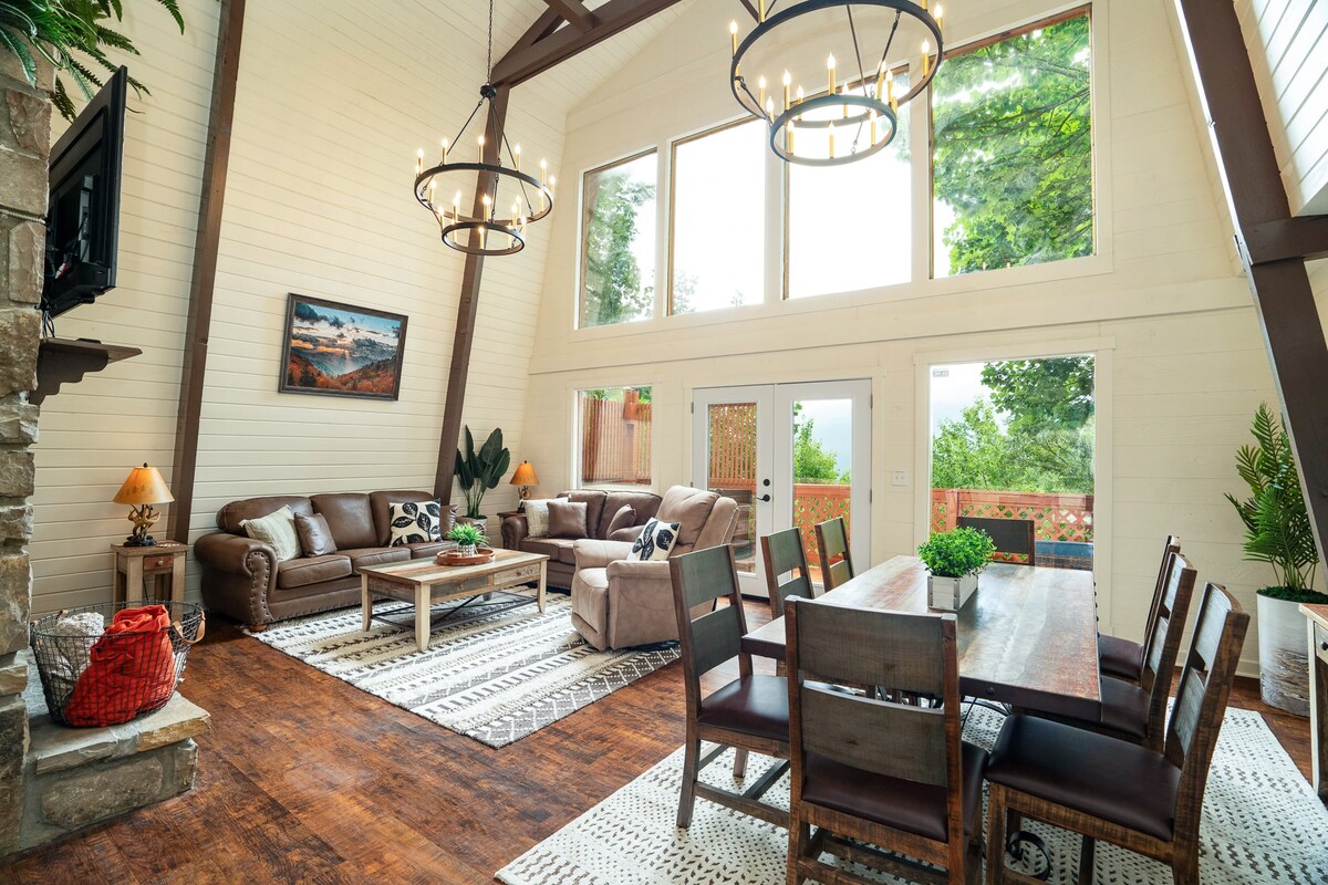 A spacious living area is characterized by high ceilings and large windows allowing natural light. The room features comfortable seating arrangements, a dining table for eight, and a decorative area rug. A stone fireplace adds warmth, while plants enhance the inviting environment.