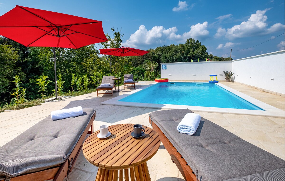 A pool area is highlighted by two sun loungers with thick cushions, set beside a pristine swimming pool. Large red umbrellas provide shade, and a wooden table holds two cups and a rolled towel. Lush greenery frames the perimeter of the space.