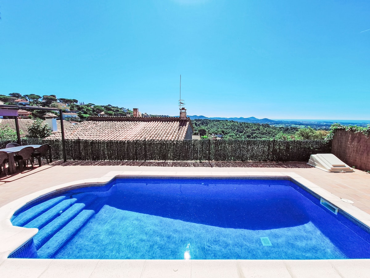 A private swimming pool features a stair entry and is surrounded by a spacious terrace. Lush trees and distant hills can be seen in the background, with views extending toward the sea under a clear blue sky.