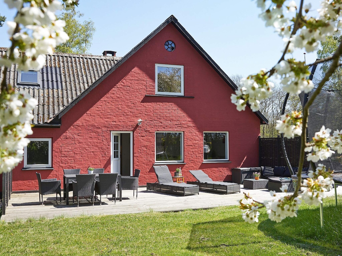A red two-story holiday home is set against a green lawn, framed by blossoming trees. The outdoor space features a wooden deck with loungers and a dining area, as well as large windows that invite natural light into the interior.
