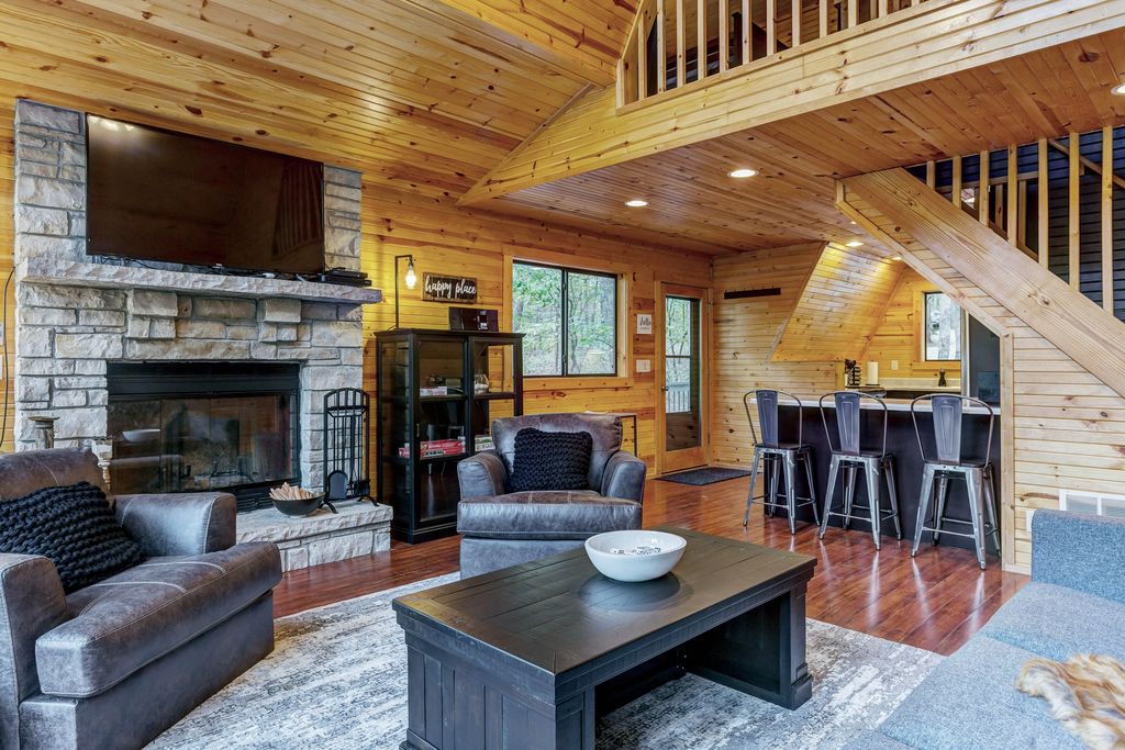 The living room features wooden walls and a cozy seating arrangement with two plush chairs and a spacious sofa. A stone fireplace with a mounted television is the focal point, while a dark coffee table sits in the center. The open layout leads to a kitchen area with bar seating.