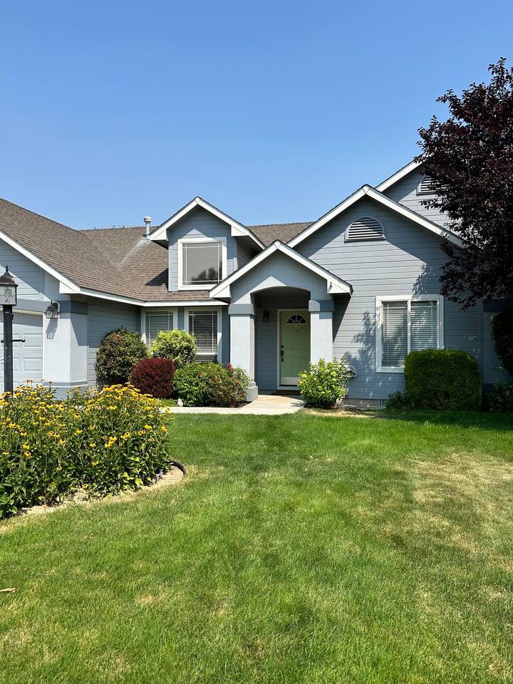 The exterior of a light blue house is highlighted, featuring a large front lawn with green grass and colorful flowers. A welcoming entryway with a green door is flanked by neatly trimmed hedges and decorative landscaping. A clear blue sky completes the scene.