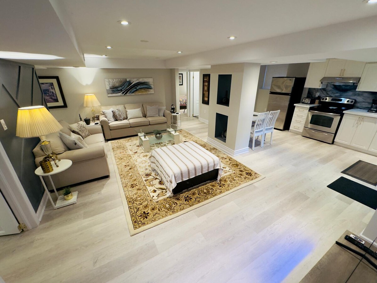 A spacious living area features a light-colored sofa and armchairs surrounding a glass coffee table. A plush area rug anchors the space, and a dining table with white chairs is positioned nearby. Modern kitchen appliances are visible, along with natural light from recessed lighting.