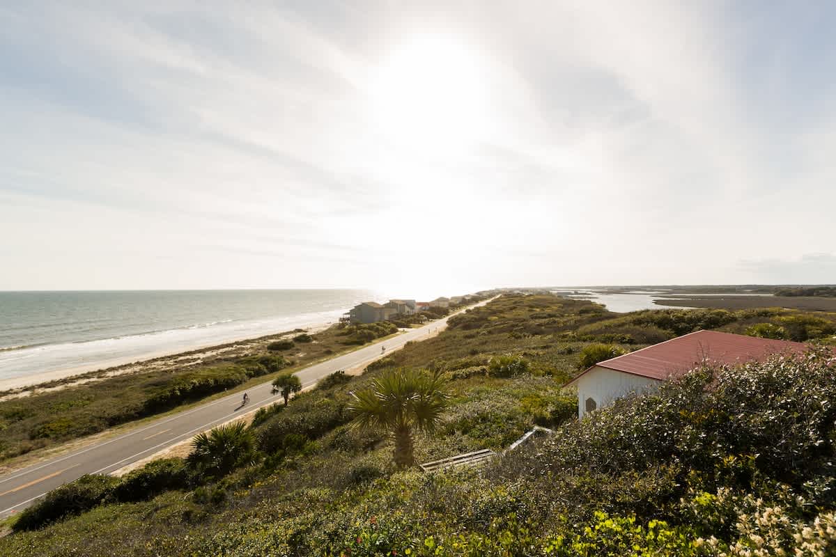 An expansive view showcases the coastline, with gentle waves lapping at sandy shores. A narrow road runs parallel to the beach, dotted with homes along the horizon. Lush greenery fills the foreground, under a bright sky that transitions from soft clouds to sunlight.