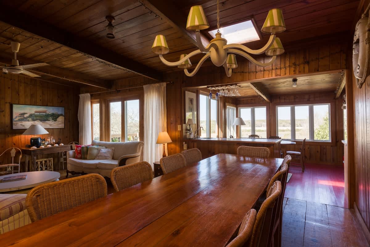 A spacious dining area features a large wooden table surrounded by wicker chairs, illuminated by a central chandelier. Natural light floods the room through multiple windows, complementing the warm knotty pine walls. The adjoining living area includes a cozy sofa, enhancing the inviting layout.